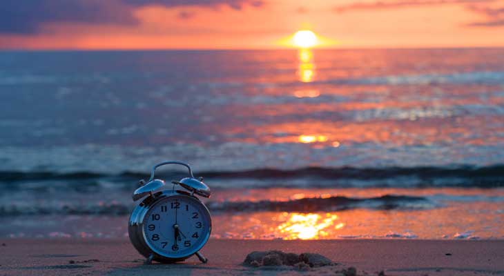 Photo of an old-style alarm clock on the beach with the sun setting