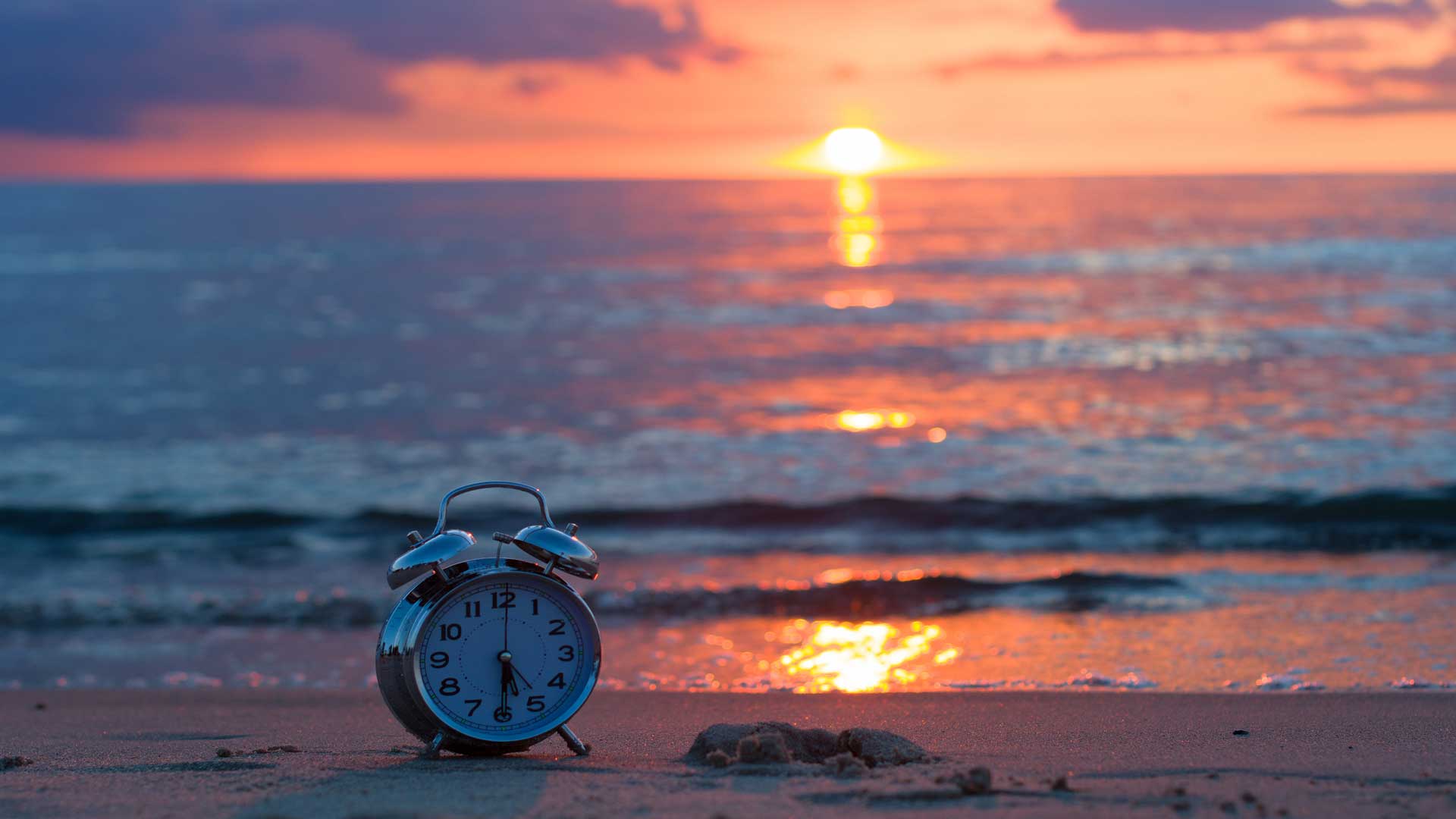 Photo of an old-style alarm clock on a beach with the sun setting